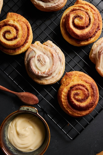 Free photo vertical shot of delicious cinnamon rolls with a metal bowl of  white glaze on a black table