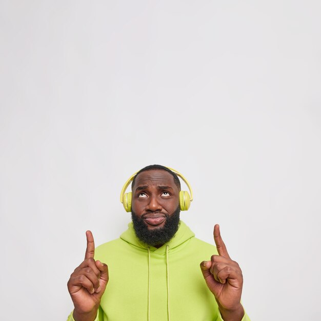 Free photo vertical shot of displeased bearded man with dark skin points index fingers above feels discontent wears wireless headphones on ears casual hoodie poses against white wall