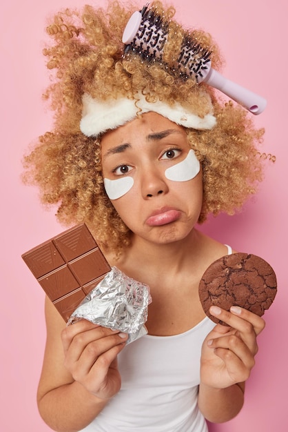 Free photo vertical shot of displeased curly haired woman holds bar of chocolate and cookie feels upset applies beauty patches under eyes feels temptation to eat delicious sweet food isolated on pink wall