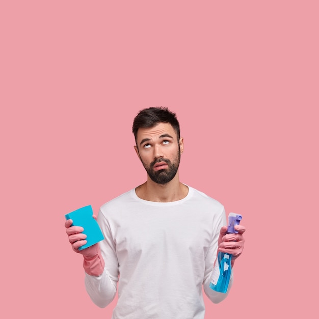 Vertical shot of displeased unshaven man focused above with tired expression, notices dirty ceiling in kitchen, wears rubber gloves for cleaning