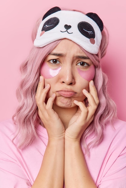 Vertical shot of dissatisfied young Asian woman has gloomy expression purses lips feels unhappy applies beauty patches under eyes wears sleepmask casual t shirt isolated over pink background