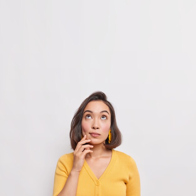 Vertical shot of dreamy thoughtful young Asian woman with dark hair focused above considers something wears casual yellow jumper isolated over white wall copy space for your advertisement