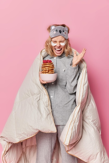 Free photo vertical shot of emotional young woman dressed in nightwear exclaims loudly keeps palm raised poses with delicious pancakes covered with blanket poses against pink background good morning concept