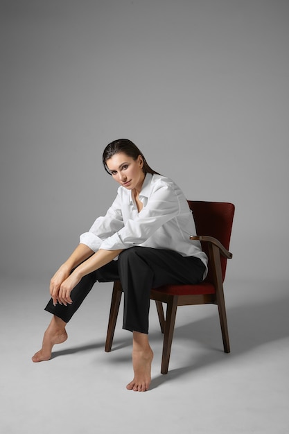 Вертикальный снимок модной привлекательной молодой брюнетки-бизнес-леди в белой рубашке и брюках, сидящей босиком в удобном кресле в расслабленной позе и отдыхающей после тяжелой работы