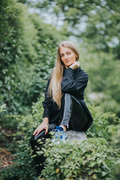 Vertical shot of a fashionable caucasian blonde female posing surrounded by greenery