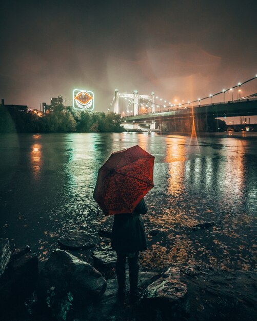 Vertical shot of a female holding a red umbrella standing near a lake in the city during nighttime