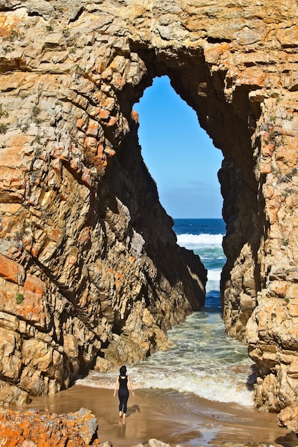 Vertical shot of a female standing in front of a cave leading to the ocean