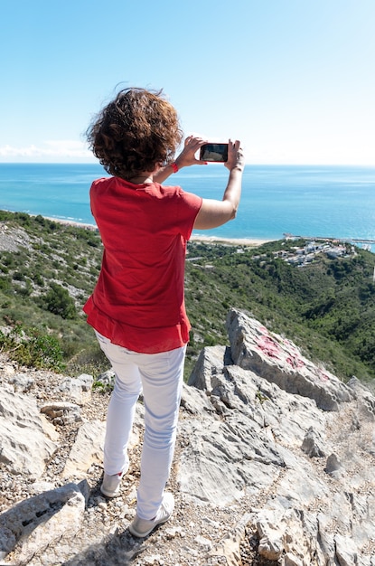Вертикальный снимок женщины, стоящей на скалах и фотографирующей красивое море