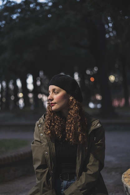 Vertical shot of a female wearing a khaki jacket and a black warm hat sitting in the park