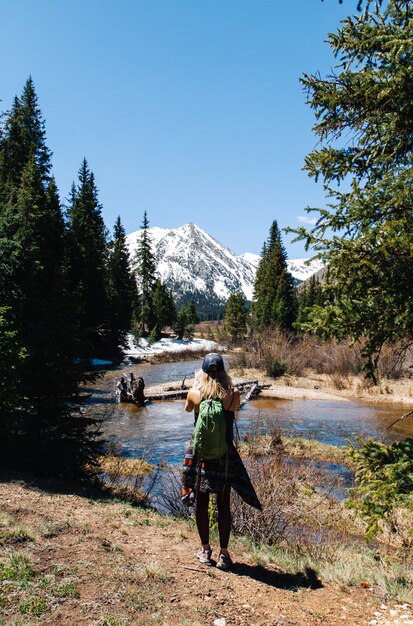 Vertical shot of a female with backpack standing near water and trees with a mountain in background