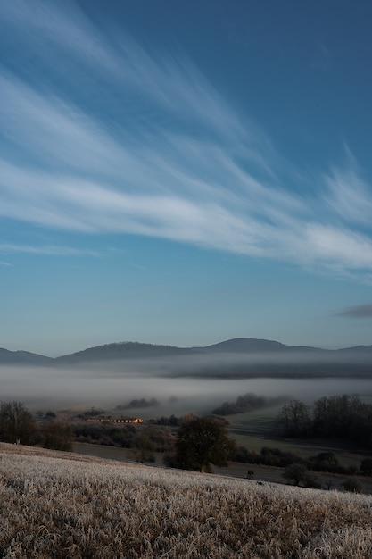 Vertical shot of a foggy field and mountains with a blue sky in the background