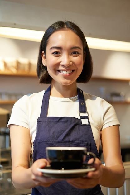 Colpo verticale di una ragazza asiatica amichevole sorridente che serve il barista del caffè che ti dà una tazza di caffè preparata