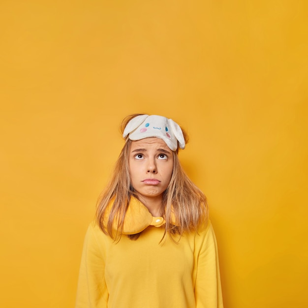 Vertical shot of frustrated sleepy woman looks above unhappily wears sleepmask neck pillow and casual jumper cannot fall asleep has sleep disorder isolated over yellow background copy space.