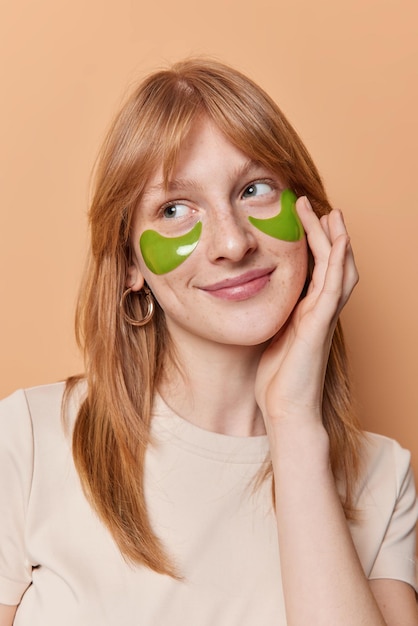 Vertical shot of gentle dreamy teenage girl touches face takes care of skin applies green nourishing patches under eyes has natural red hair dressed in casual t shirt isolated over beige wall