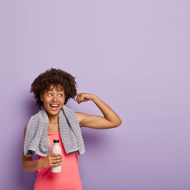 Vertical shot of glad dark skinned female raises muscular arm, has fitness workout, drinks water from bottle