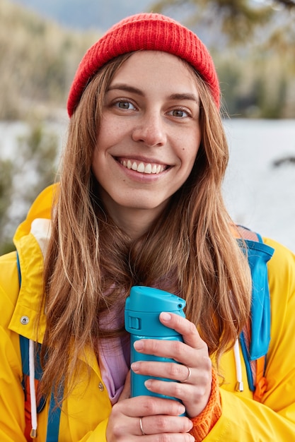 Vertical shot of glad smiling woman wears red headgear, yellow raincoat, warms herself with hot drink from flask