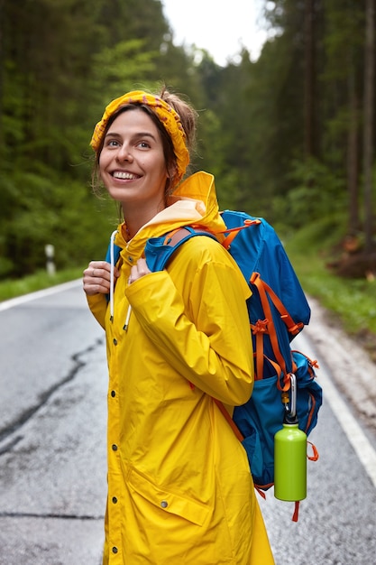 Вертикальный снимок довольной улыбающейся молодой европейской женщины в желтой повязке на голову, плаще и рюкзаке