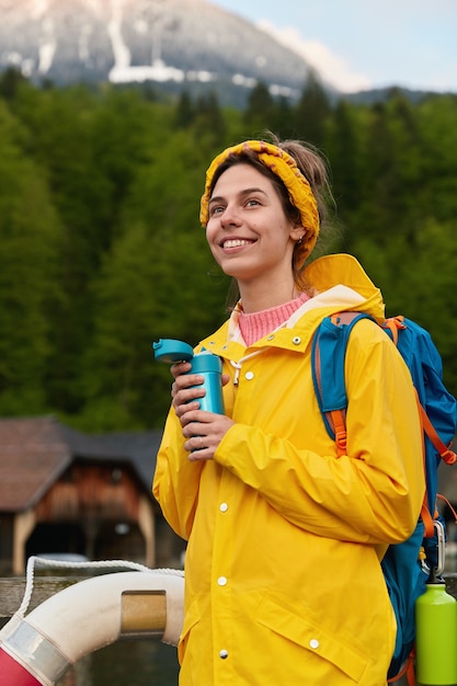 Vertical shot of glad young European female in yellow anorak with rucksack, poses at raft boat against rocky mountains and forest