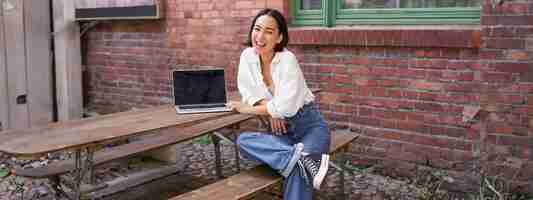 Free photo vertical shot of goodlooking asian woman with laptop sitting in outdoor cafe drinking coffee and