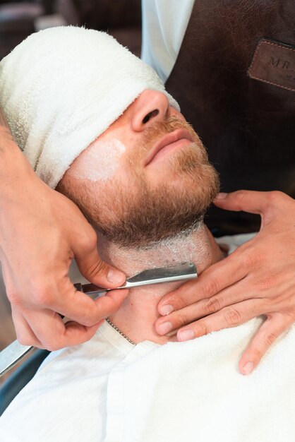 Vertical shot of a hairdresser shaving the beard of a young customer in a towel with a blade