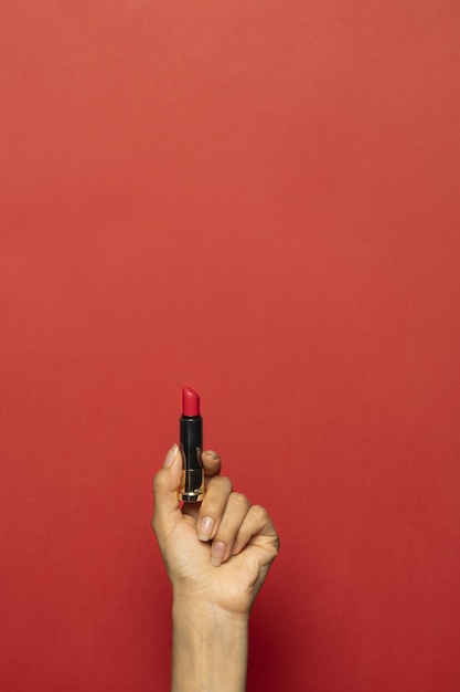 Free photo vertical shot of a hand holding a lipstick isolated on a red wall