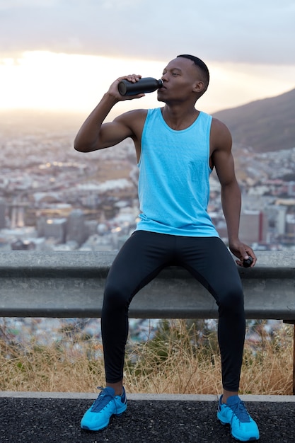 Vertical shot of handsome African American man in sportswear drinks water after fitness training, refreshes with beverage, poses over mountain hill, feels fatigue. Sport and rejuvenation concept