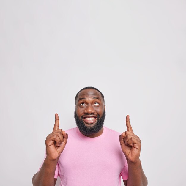 Vertical shot of handsome bearded adult man has thick beard points above with both index fingers shows space for your advertising content look happily overhead isolated over grey wall