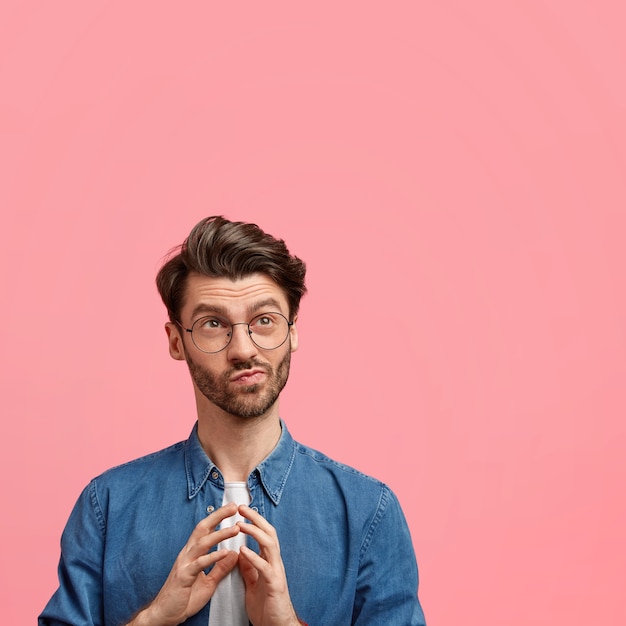 Vertical shot of handsome pensive unshaven young male, keeps hands pressed together, looks pensively upwards, dressed in elegant denim shirt, isolated over pink wall with copy space aside