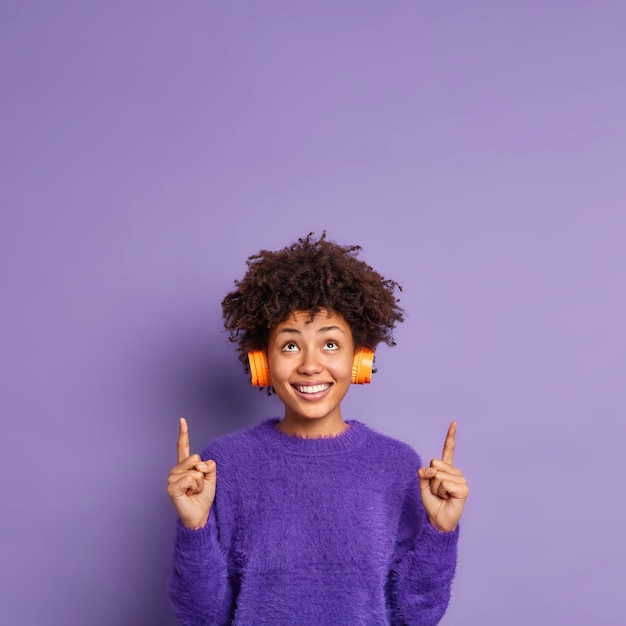 Colpo verticale della donna afroamericana felice in ponticello casuale indica sopra sullo spazio della copia ha sorrisi di espressione del viso positivo indossa volentieri cuffie stereo sulle orecchie