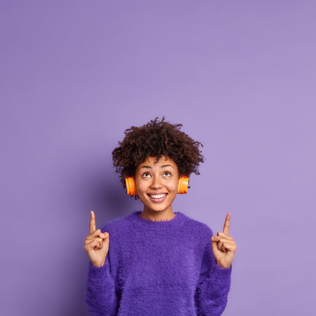 Vertical shot of happy African American woman in casual jumper indicates above on copy space has positive face expression smiles gladfully wears stereo headphones on ears 