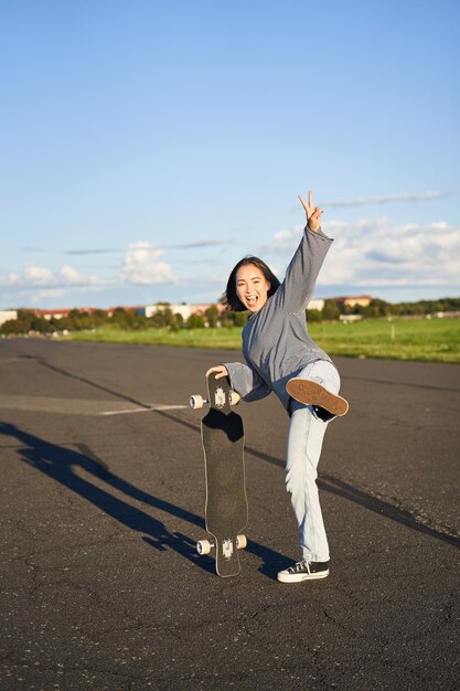 スケートボードに立ってジャンプし、スケートをしている笑顔の女性の幸せなアジアのスケーターの女の子の垂直ショット