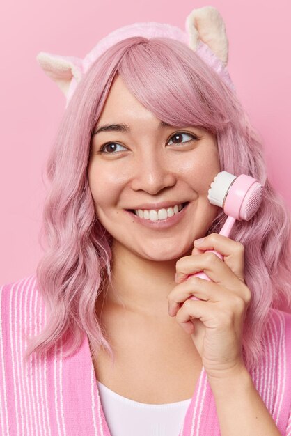 Vertical shot of happy Asian woman with pink dyed long hair uses facial cleansing brush takes care of skin and complexion wears headband concentrated away poses indoor. Facial beauty treatment.