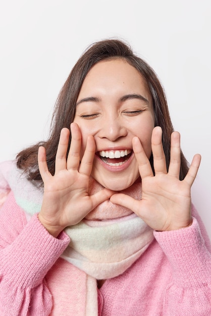 Il colpo verticale di una donna felice e spensierata divertito tiene le mani vicino alla bocca gli occhi chiusi si sente sussurra gioiosa qualcosa indossa un maglione e un maglione caldo isolato su sfondo bianco. concetto di emozioni