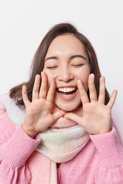 Vertical shot of happy carefree amused woman keeps hands near mouth eyes closed feels joyful whispers something wears sweater and warm sweater isolated over white background. Emotions concept