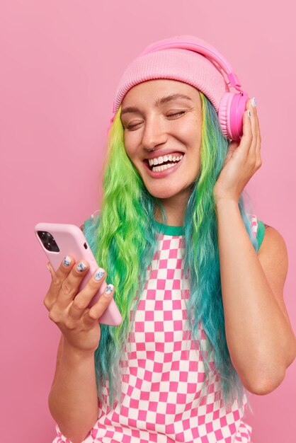 Вертикальный снимок счастливого беззаботного подростка с длинными яркими волосами, выбирающего любимую песню из плейлиста, держит мобильный телефон в беспроводных наушниках, в шляпе и платье, изолированном на розовом