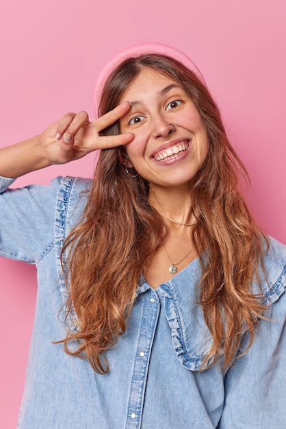 Il colpo verticale di giovane donna europea spensierata felice con i capelli lunghi fa il gesto di pace sui sorrisi degli occhi ampiamente vestito con camicia di jeans e fascia isolata su sfondo rosa. segno della discoteca.