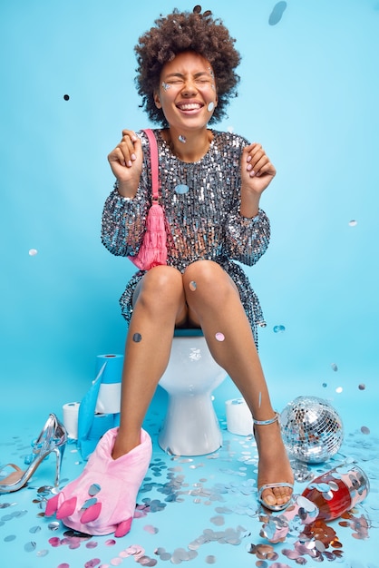Вертикальный снимок счастливой кудрявой афроамериканки, сжимающей кулаки, празднует, что кто-то носит праздничную одежду, позирует на унитазе в грязном туалете после вечеринки, веселится в приподнятом настроении