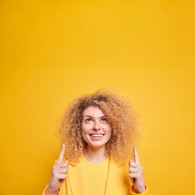 Vertical shot of happy curly haired woman points finger up shows amazing offer overhead smiles toothily 