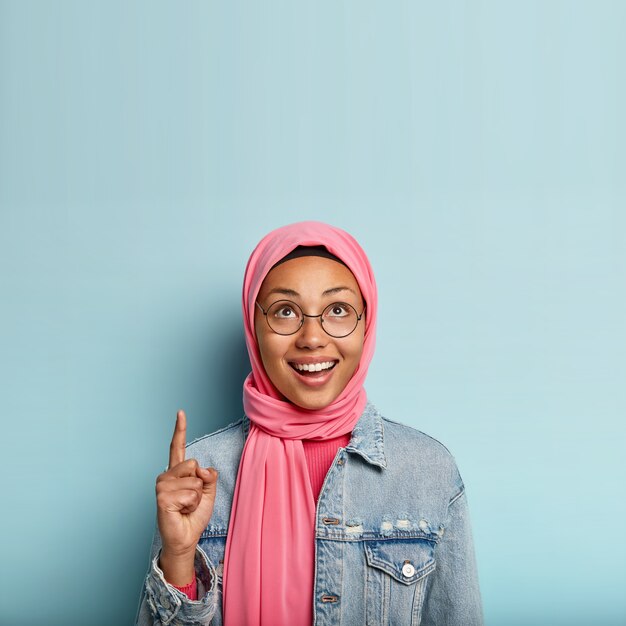 Vertical shot of happy dark skinned female Muslim woman poses over blue space