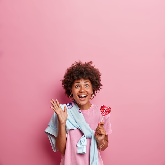 Vertical shot of happy dark skinned girl looks above with glad face expression, smiles broadly and raises hand, holds delicious lollipop, sees something amazing upwards, isolated on pink wall