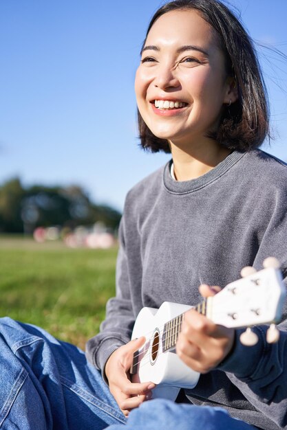 Vertical shot of happy korean girl sitting in park learning how to play ukulele singing and relaxing