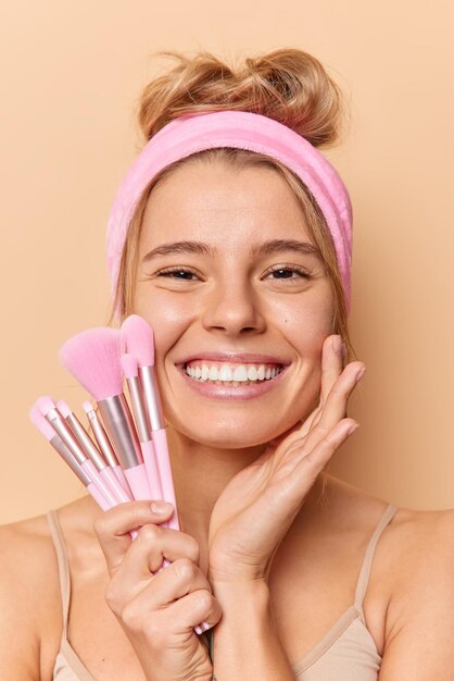 Vertical shot of happy positive young woman touches face gently smiles toothily wears pink headband holds cosmetic brushes looks directly at camera isolated over beige background puts make up on