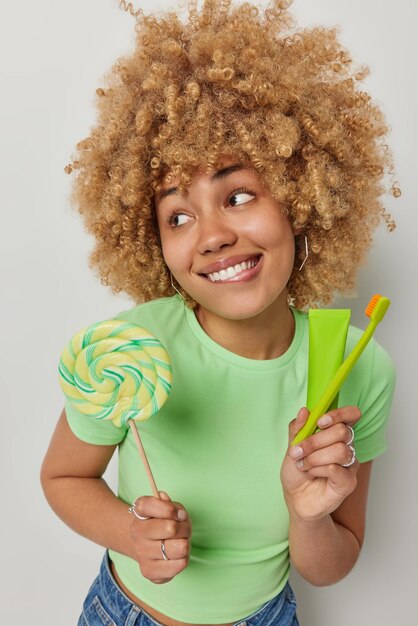 Vertical shot of happy teenage girl refuses to eat lollipop beacause wants to have healthy teeth holds tube of toothpaste and toothbrush wears casual green t shirt isolated over white background