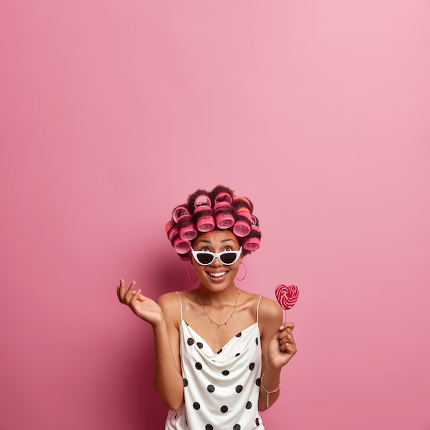 Colpo verticale di donna felice concentrata sopra, alza la mano e tiene il lecca-lecca, indossa bigodini e fa una bella acconciatura, si prende cura dei capelli, vestito con abito a pois e occhiali da sole