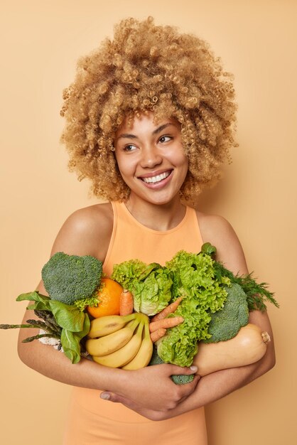 Vertical shot of happy woman poses with fresh green vegetables and fruits keeps to healthy diet wears casual t shirt isolated over brown background Homegrown grocery Vegeterian food concept