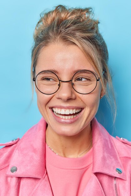 Vertical shot of happy woman with healthy skin toothy smile looks away gladfully notices something funny wears round transparent spectacles pink jacket isolated over blue background Happiness