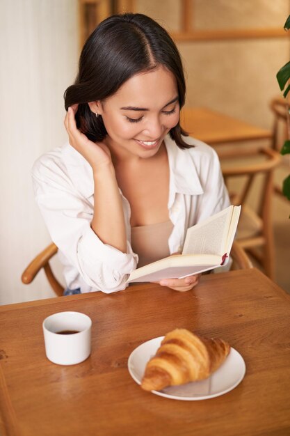 幸せな若いアジア女性の垂直ショットは、カフェでコーヒーを飲みながら本を読んで楽しんでいます。