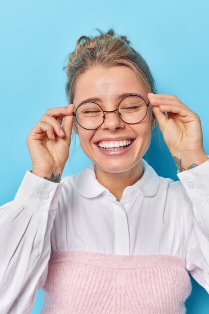 幸せな若い白人女性の垂直ショットは、喜びの笑顔から目を閉じ、青い背景に白いシャツのポーズを着た眼鏡の縁に歯を見せて手を保ちます。ポジティブな感情の概念