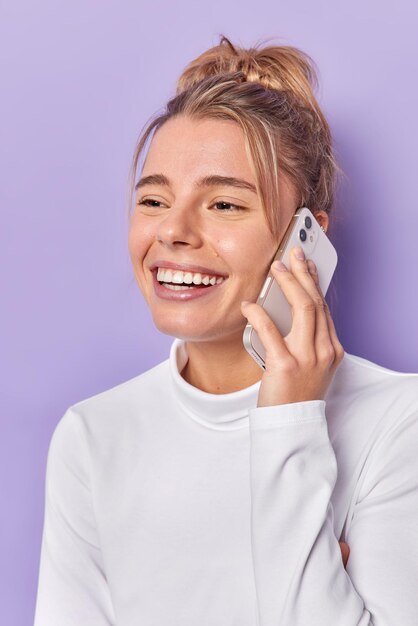 Вертикальный снимок счастливой молодой женщины, которая разговаривает по мобильному телефону с улыбкой лучшего друга, с удовольствием наслаждается приятным разговором, одетая в повседневный белый джемпер на фиолетовом фоне. Коммуникация
