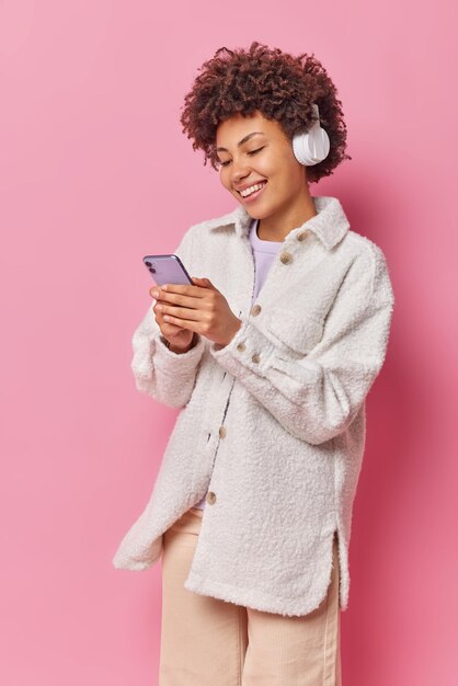 Vertical shot of joyful woman with curly hair holds mobile phone chats online and listens favorite music in headphones wears stylish casual clothes isolated over pink wall